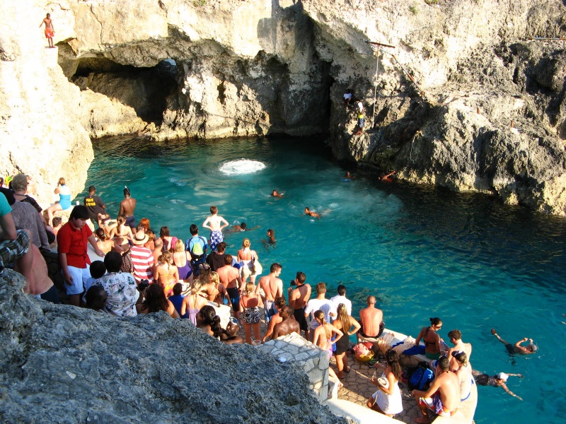 a crowd of people standing and sitting on a rock ledge that's been changed to be a balcony. The ocean, with swimmers in it, is below them.