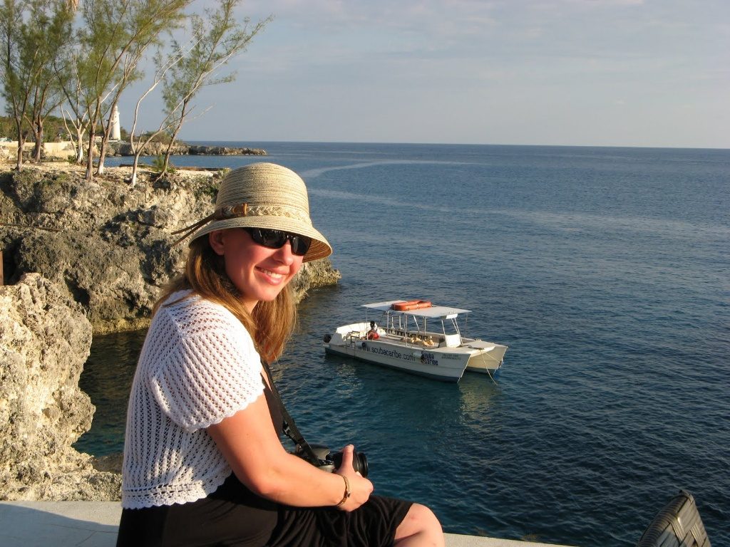 This is me, wearing a tan hat with a small brim, sunglasses, black dress, and white short-sleeved shrug. I'm sitting on a rock, overlooking the ocean and there is a boat in the distance. It was taken at Rick's Cafe in Negril, Jamaica.