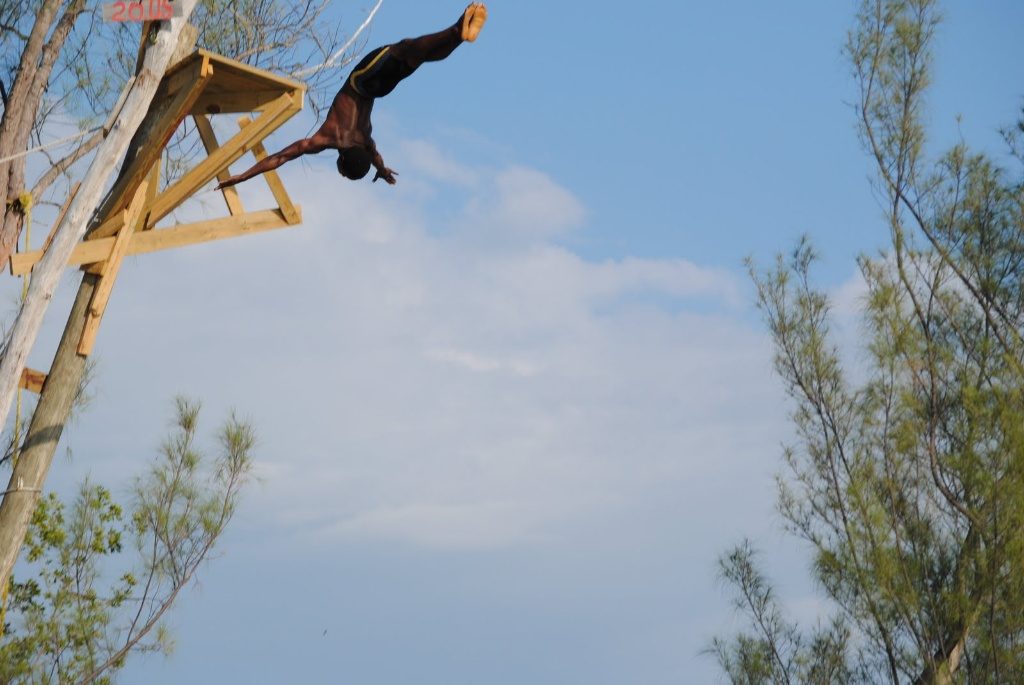 A young Jamaican man doing an incredible dive off a high platform. His head is down and his feet are in the air. 