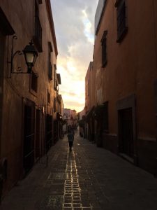 Moving to Mexico: A picture of a long cobblestone style walkway between two buildings, around sunset. Clouds are blocking the sun. 