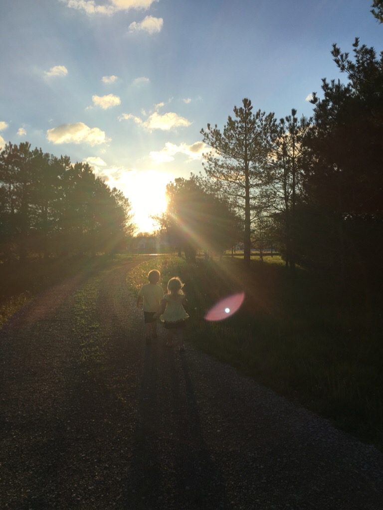 Two children, holding hands, walk into the sunset down a gravel driveway. 