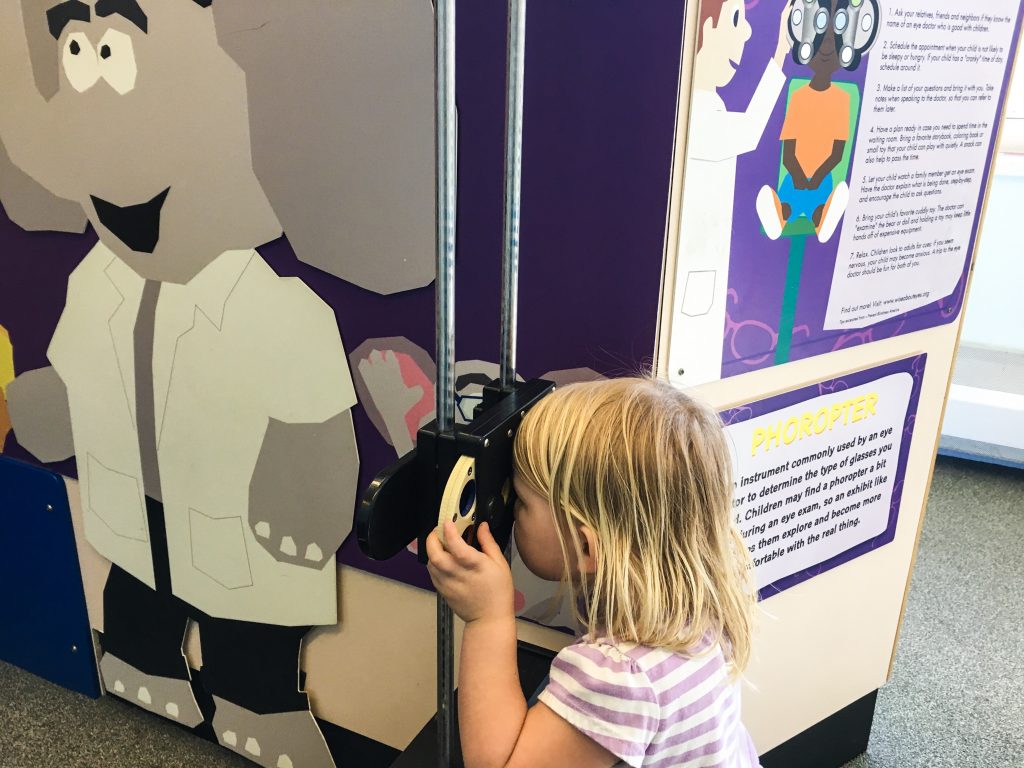 A blonde girl looking through child sized eye doctor eyesight checker thing. Columbus children's museum ohio