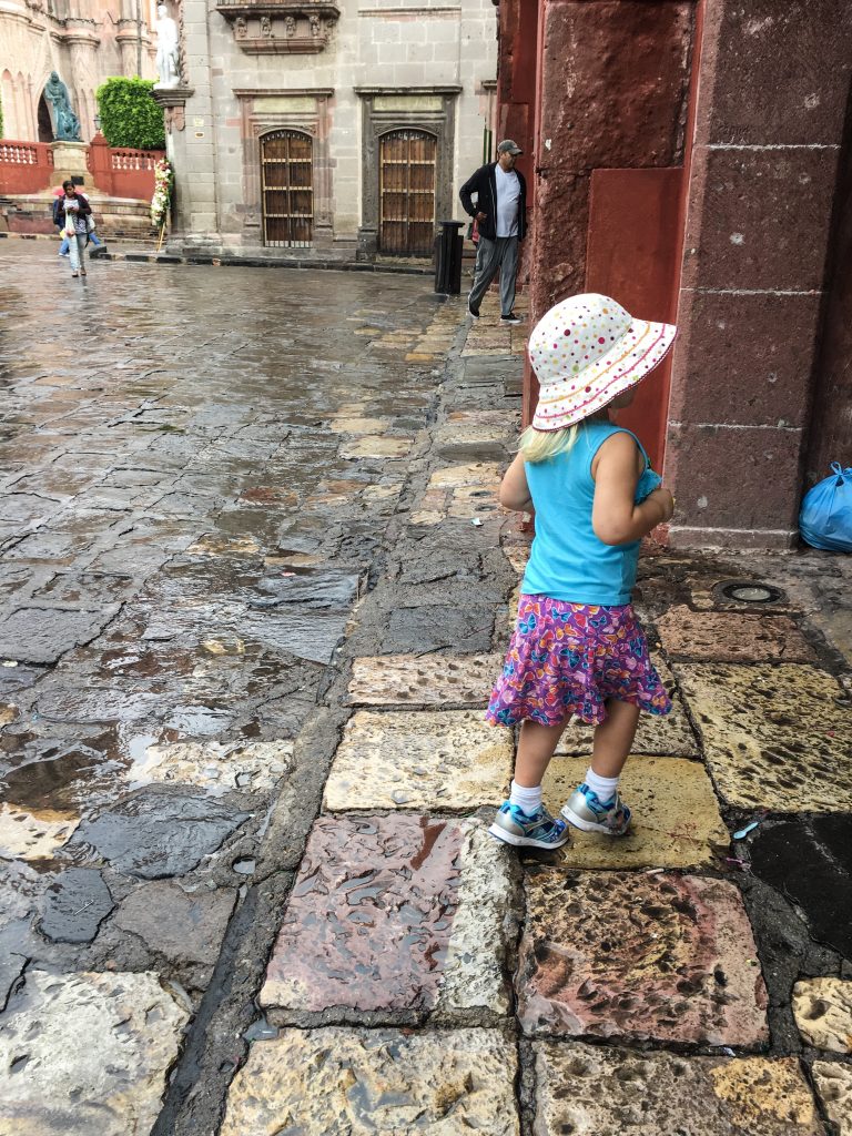 pictured: a litle girl in a hat wandering in the overcast, rainy area, surronded by old buildings and stone streets. 