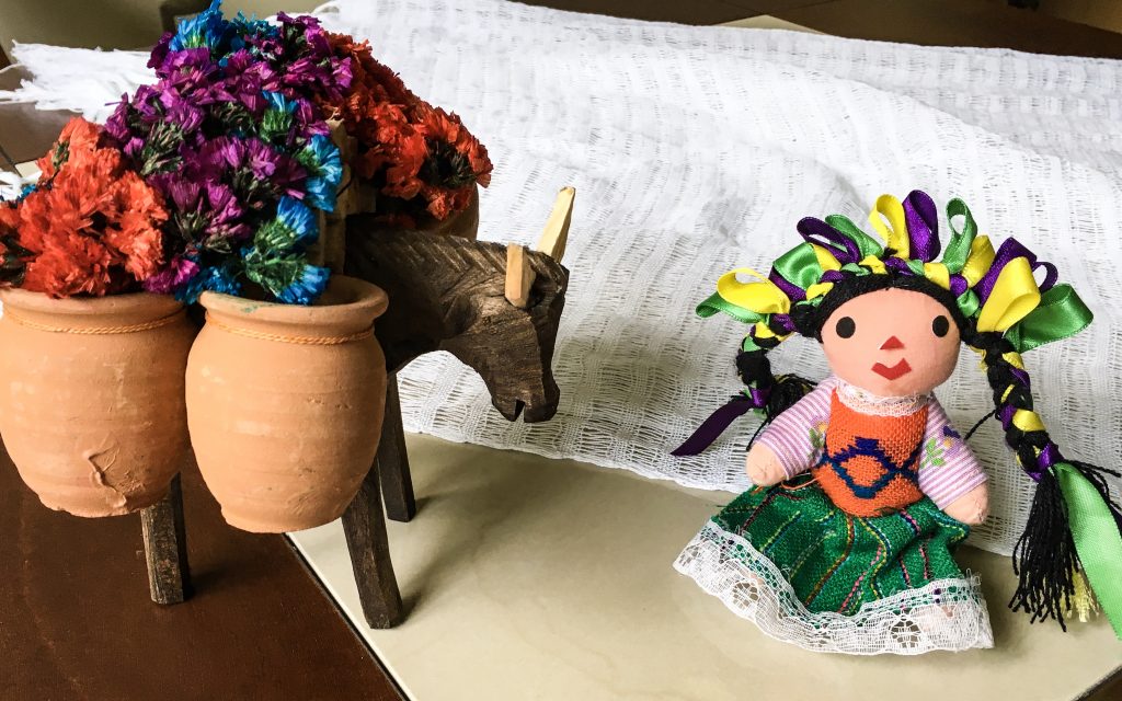 A wooden donkey carrying terra-cotta pots filled with artificial flowers, a doll with green, yellow, and purple bows in her black hair, and a white scarf apparently from Oaxaca