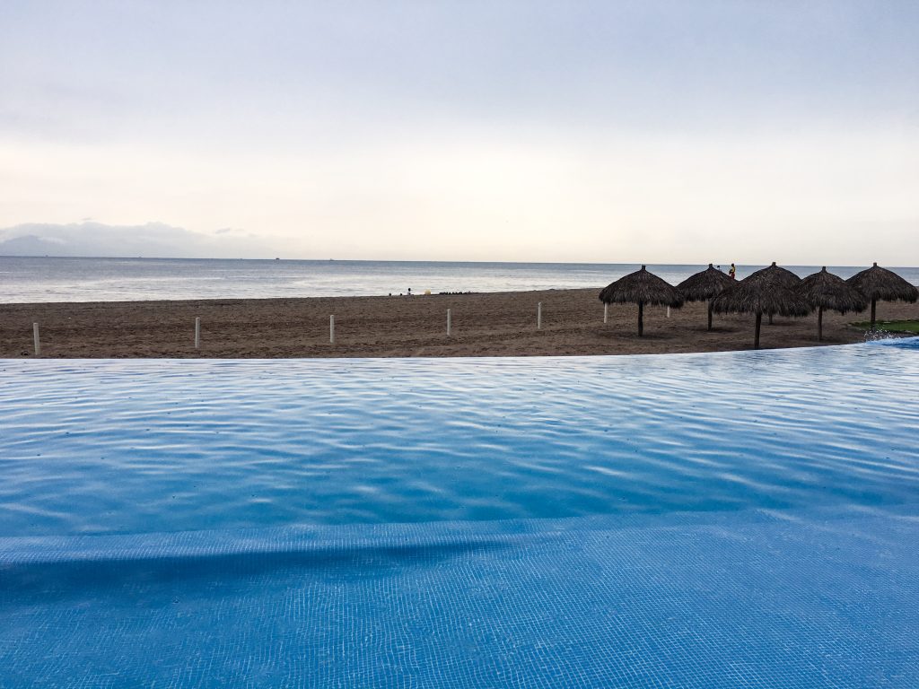 Pictured an infinity pool where it looks like the pool continues into the beach and ocean.