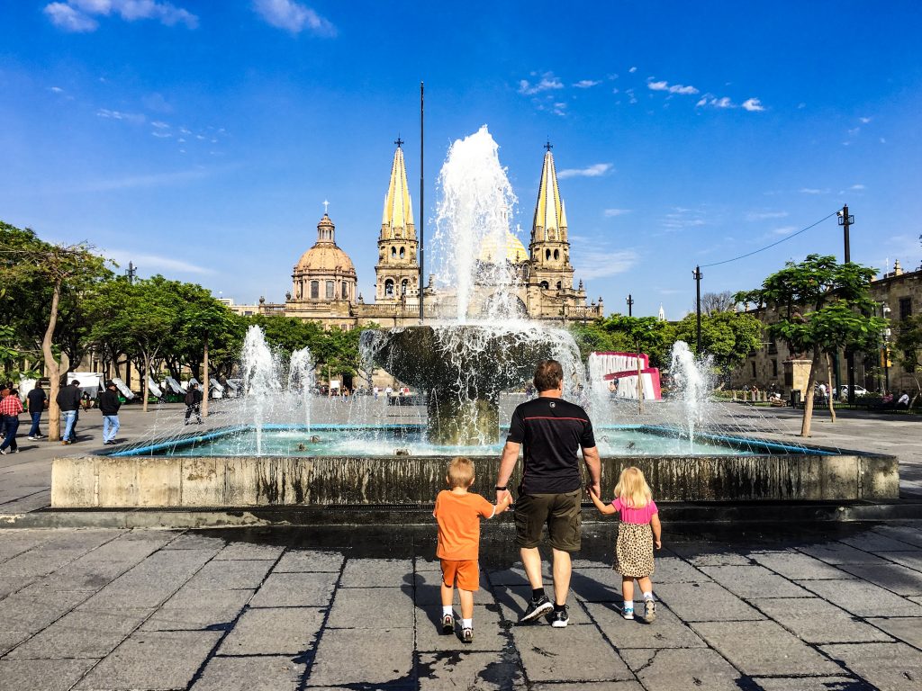 A fatherholding hands with two children, facing a fountain. There is a church and the guadalajara sign in the far background.