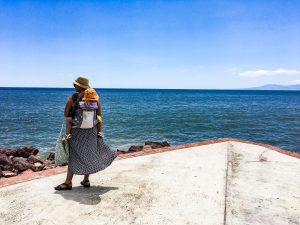 A woman faces the Pacific Ocean while wearing a child on her back in a carrier. 