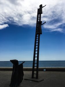 Statue: a person in a cloak reaching out, a ladder with two children in cloaks also reaching out towards the sea 