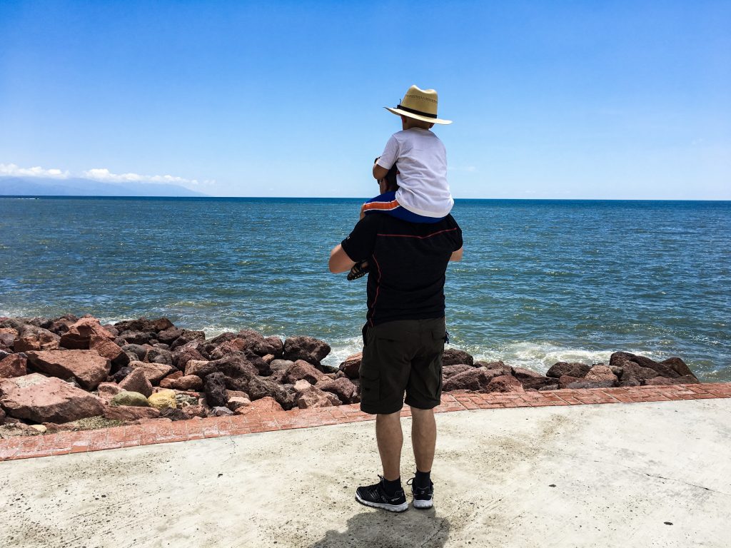 A boy sits on his father's shoulders and they are facing the Pacific Ocean.