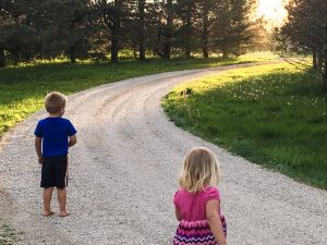 Two kids facing away from the camera on a stone driveway, as the sun sets in the background.