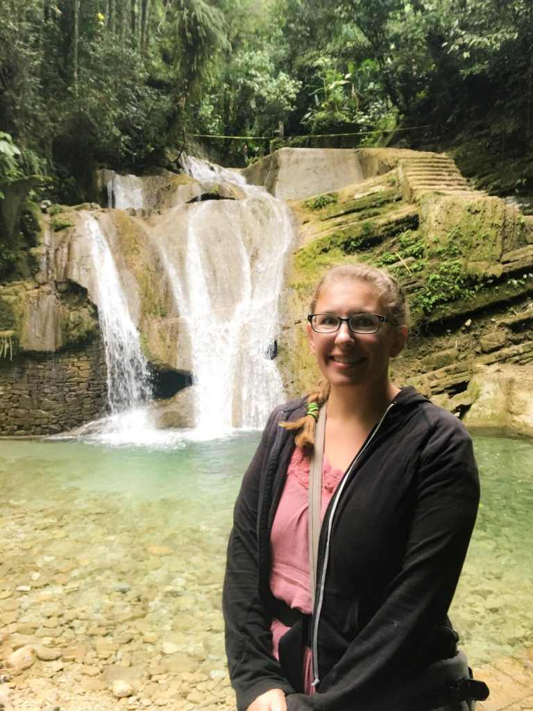 the author, wearing her brown here in a braid and glasses, stands in front of a waterfall at Las Pozas Edward James.