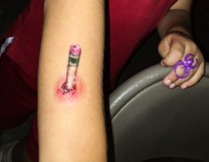 a child's arm, with a temporary tattoo on it that looks like a pencil was stabbed into the arm. in the backgound is the child's other hand wearing a purple spider ring.