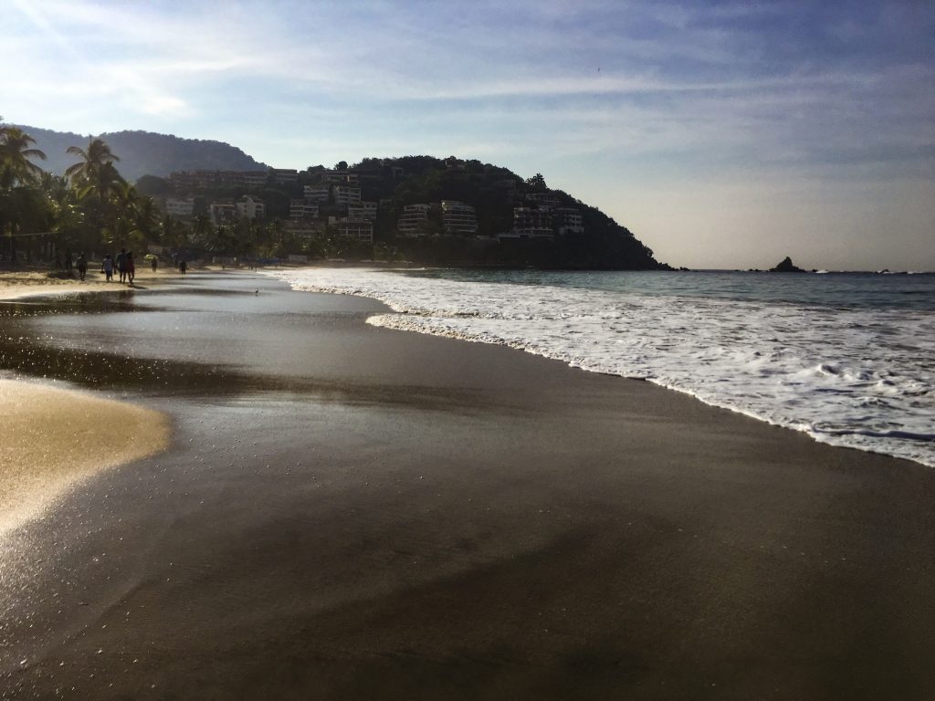 a picture of sand (foreground), the ocean on the right, and small mountains in the background.