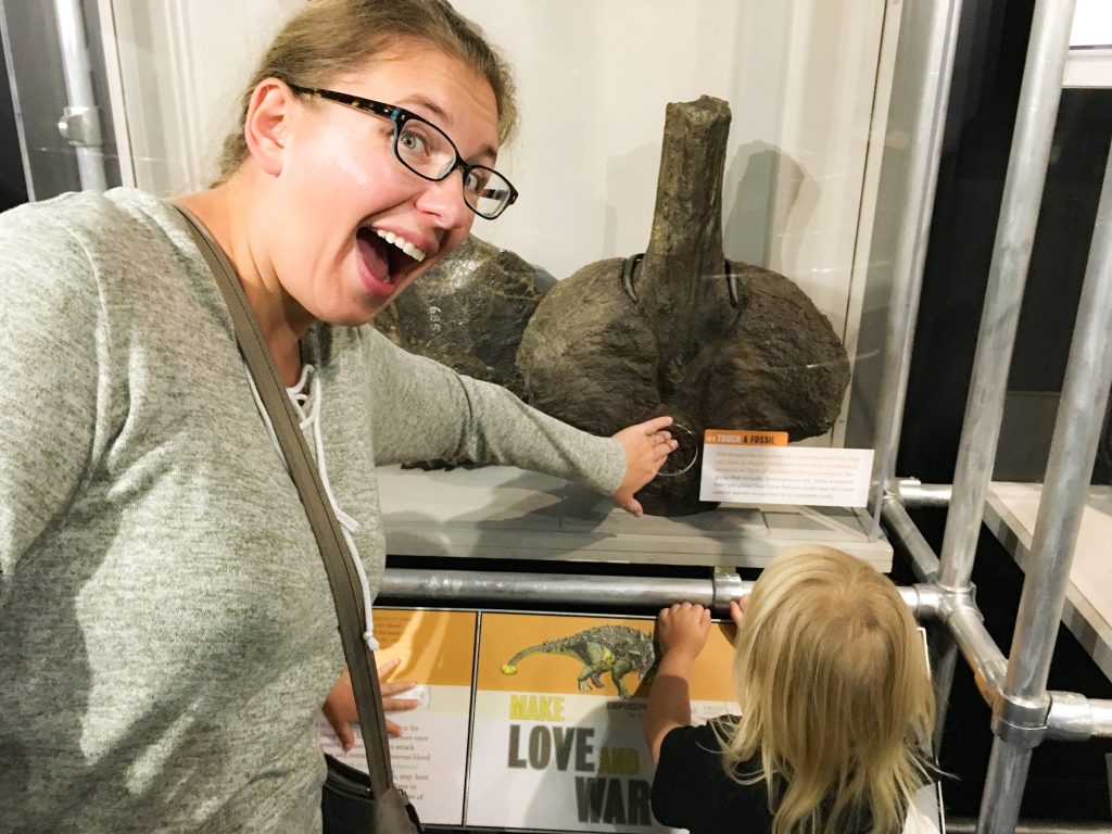 the author, a light brown haired woman wearing black glasses, with a huge smile as she touches a fossil inside a glass case.