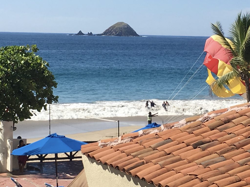 The view from my blacony. There is a roof in the right foreground and a red and yellow parachute "deflating" in the right mid ground. The beach and ocean are in the background. barcelo ixtapa beach all inclusive