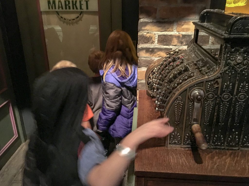 a kid wearing the hood of his coat stands at an old fashioned cash register. Three kids are in the backgound.