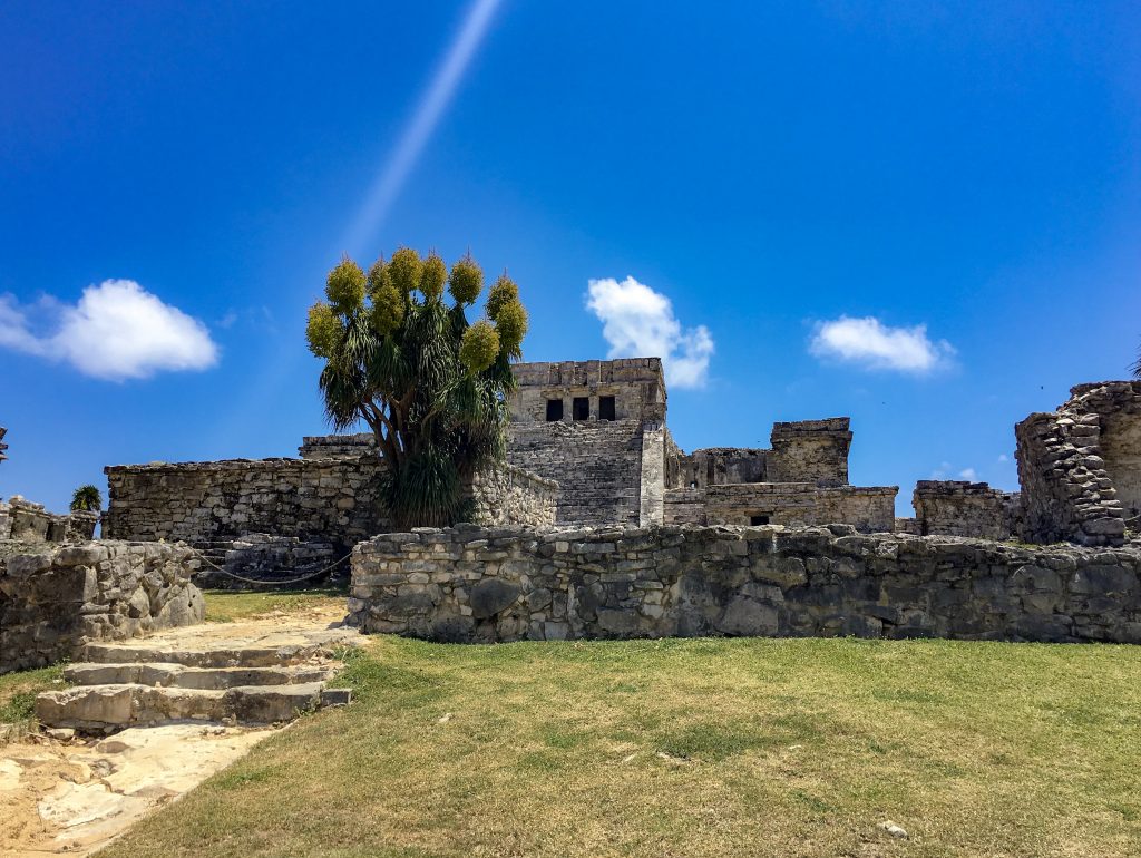 One of the standing ruins of Tulum. Slightly green grass in front, with a brilliant blue sky in the back and three small, fluffy white clouds dot the horizon.