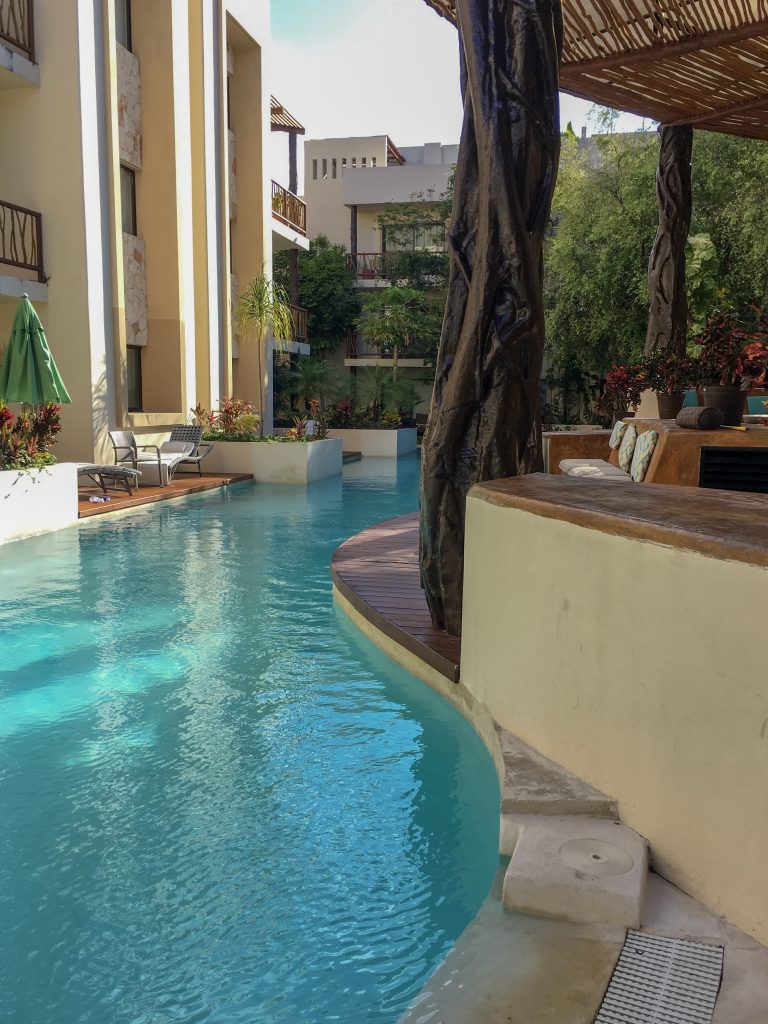 image is of a large, curved pool, surrounded by 3 story, white condo buildings. In the middle, there is a fake tree supporting a slatted roof over a seating area. The pool of our Tulum vacation!