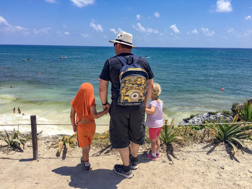 a father holding the hands of two children on either side. The child on the right is wearing orange and his shirt covers his head. The other child is wearing a purple and white striped shirt with pink shorts. All three are looking out over the ocean.