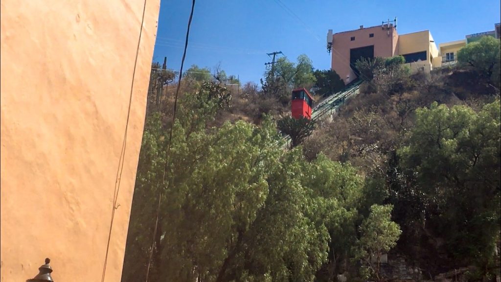 A red car on a track travels up (and down) the mountainside, whcih is covered in trees and bushes which are green.