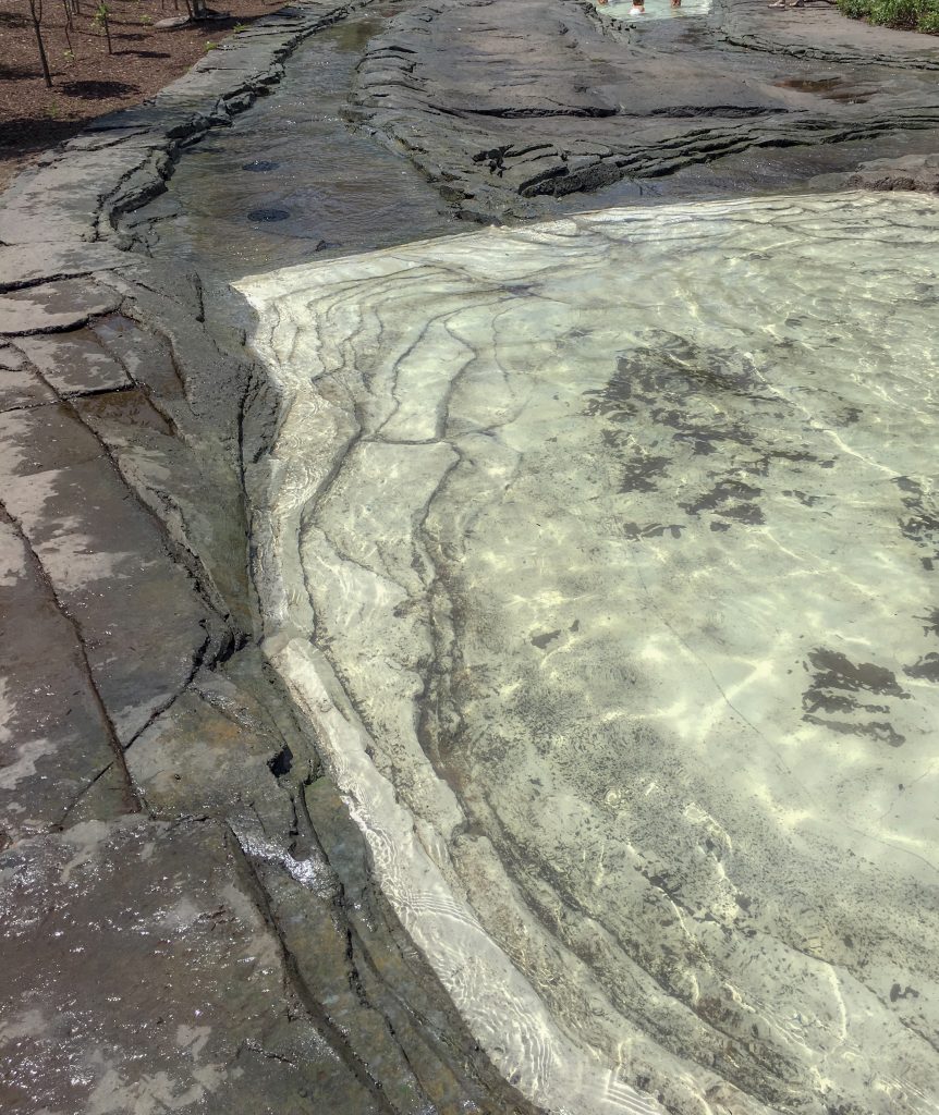 A faux creekbed, mostly gray, shale, but the bottom of the pool is lighter, almost white. A creek bed heads off in the distance. All man made.