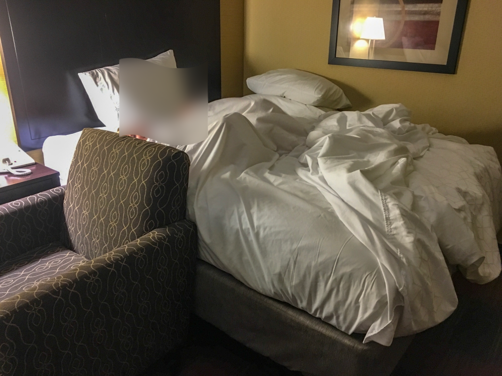 Solo parent travel means getting innovated. A hotel bed, with an armchair pushed up to it as a "railing" and pillows stacked on a luggage rack (unseen) in the background.