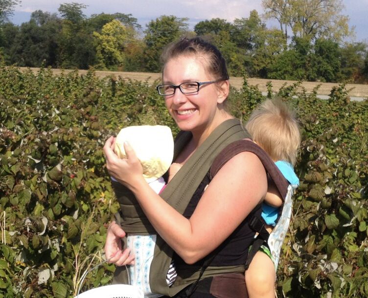 An exhausted woman with a young baby in a carrier on her front and a young toddler in a baby carrier on her back.