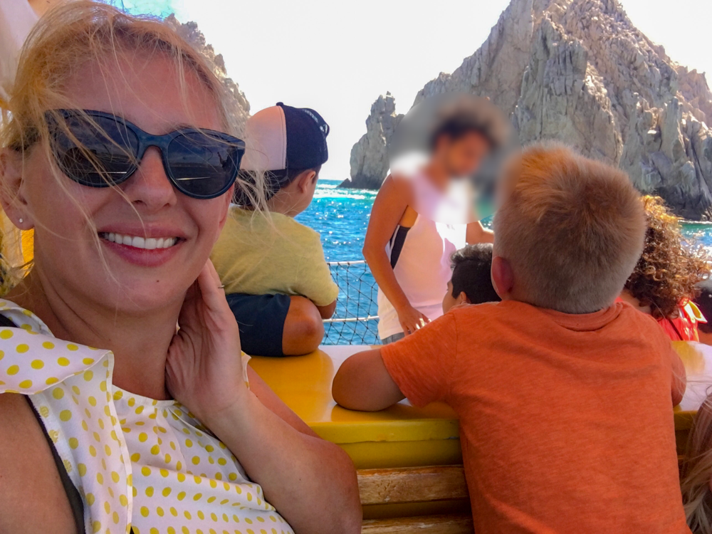 A woman wearing a white shirt with yellow polkadots, takes a selfie in front of El Arco, but a man (face blurred) is blocking most of the arch. A little boy sits to the right in an orange shirt, looking back at the arch. One of the fun things for kids to do in Cabo.