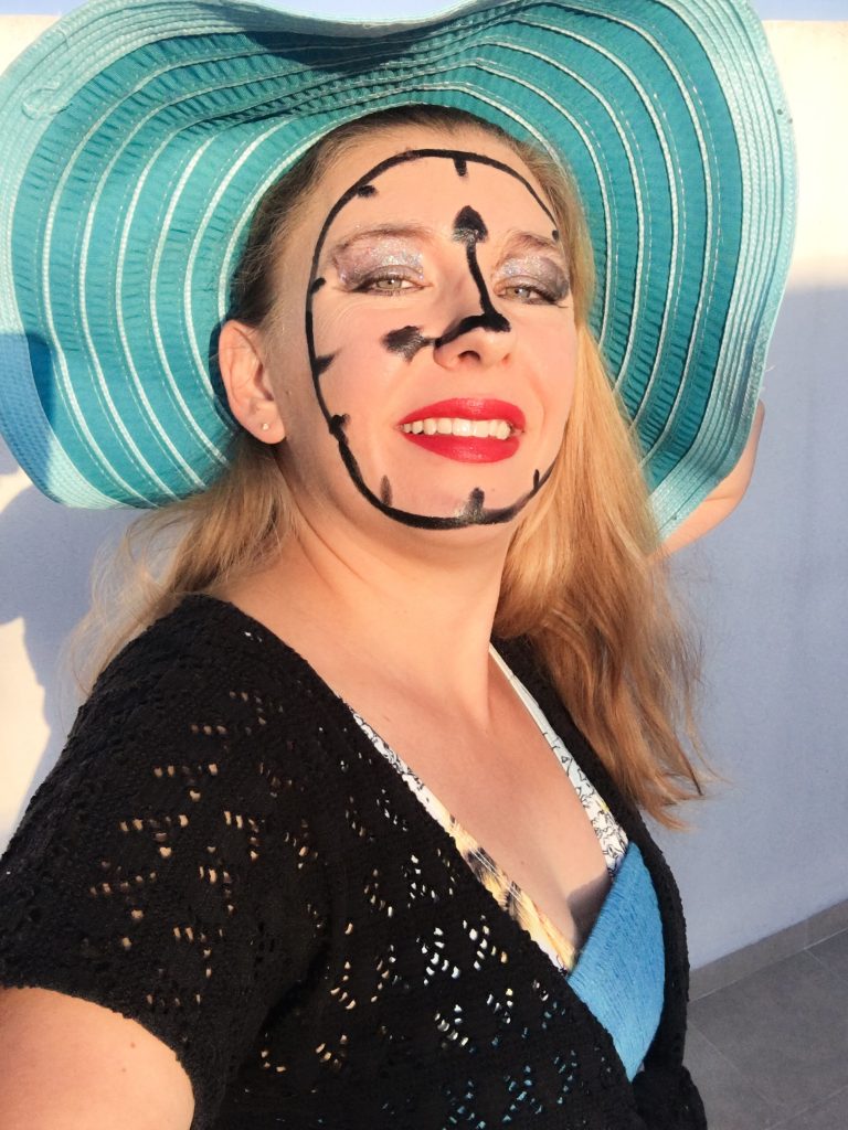 A woman posing as if she's a tourist, in a floppy sunhat, swim suit, and a clock painted on her face because she's a "time traveler"