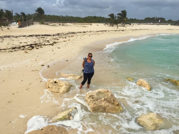 A large sandy beach dominates the majority of the photo. In the foreground, we can see the surf swirling around some sand colored boulders. A woman stands just behind them in a blue shirt and black mid-calf length pants. to the right, the turquoise sea.