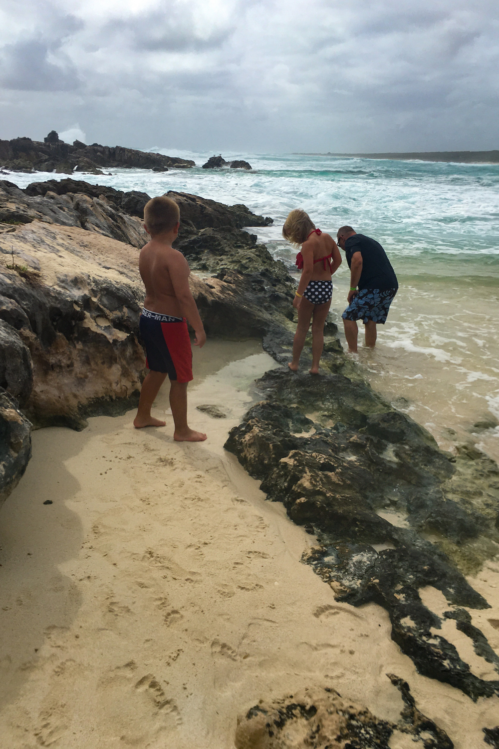 Chen Rio Cozumel, a beach mixed with rocks and plenty of sand. A family stands in the middle, all checking out the ocean as they stand on the sand. The ocean is turquoise and stunning in the background.