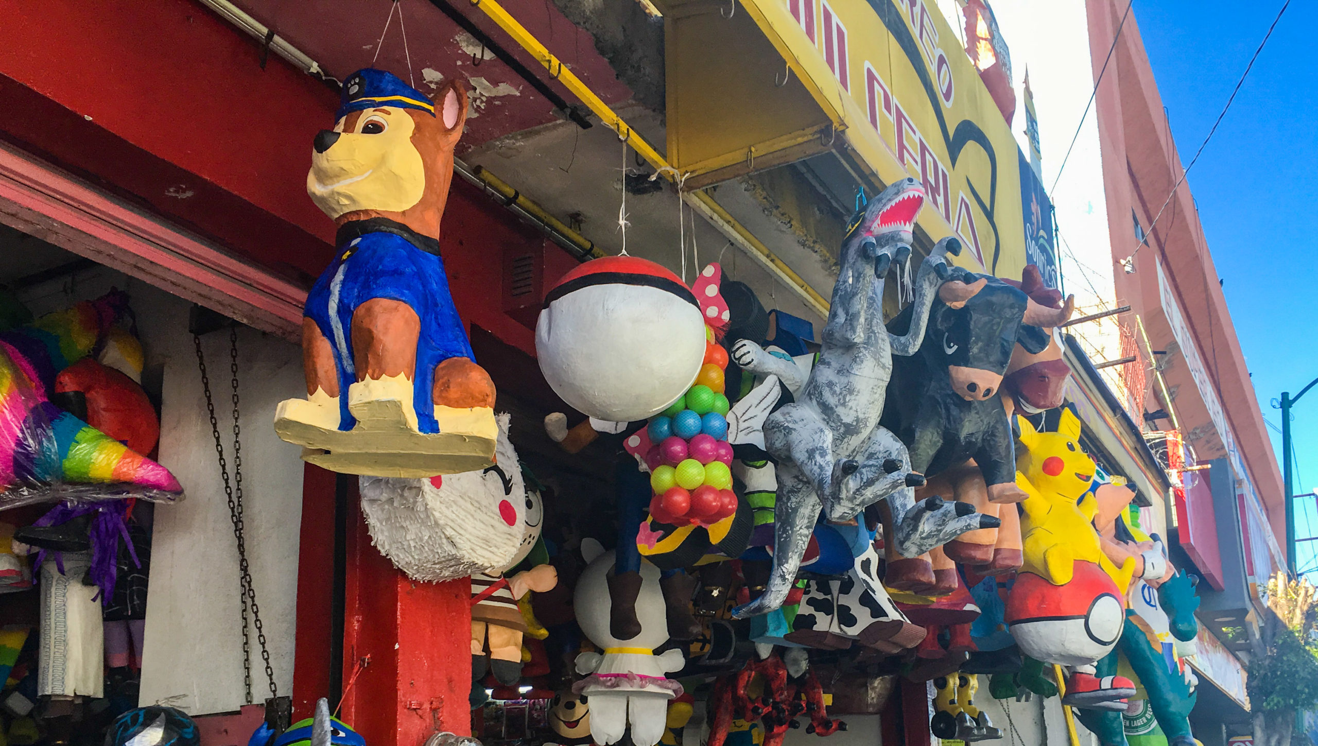 Pinatas handing from the ceiling of a shop. They include Chase from Paw Patrol, a sphere that's white on the bottom, orange on top, and a black stripe around the middle, a bull, a dinosaur, a pokemon character, and more.