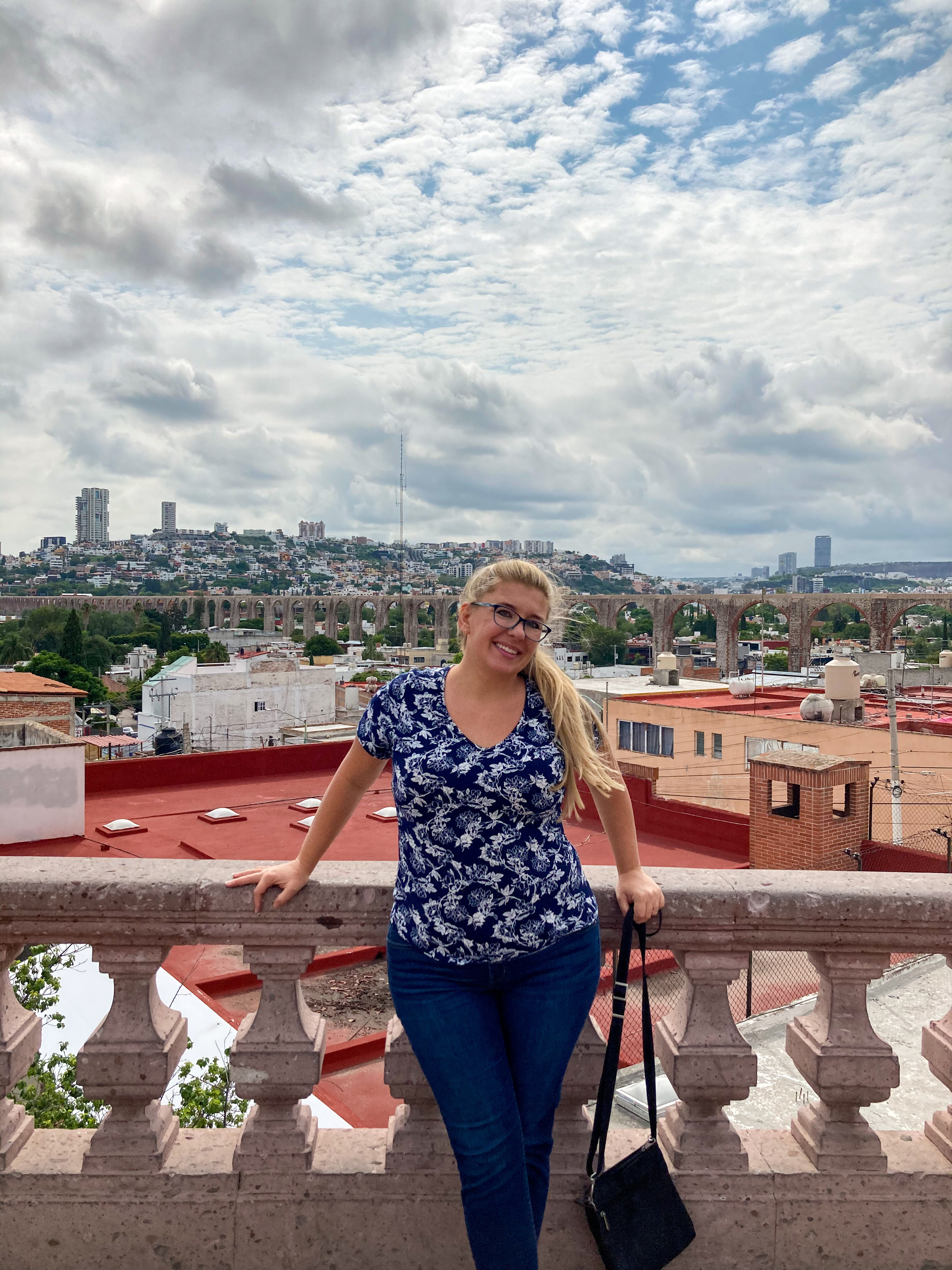 A blonde woman wearing a dark blue and white shirt with dark jeans poses in front of the aqueduct in Queretaro from becoming permanent residents in Mexico.