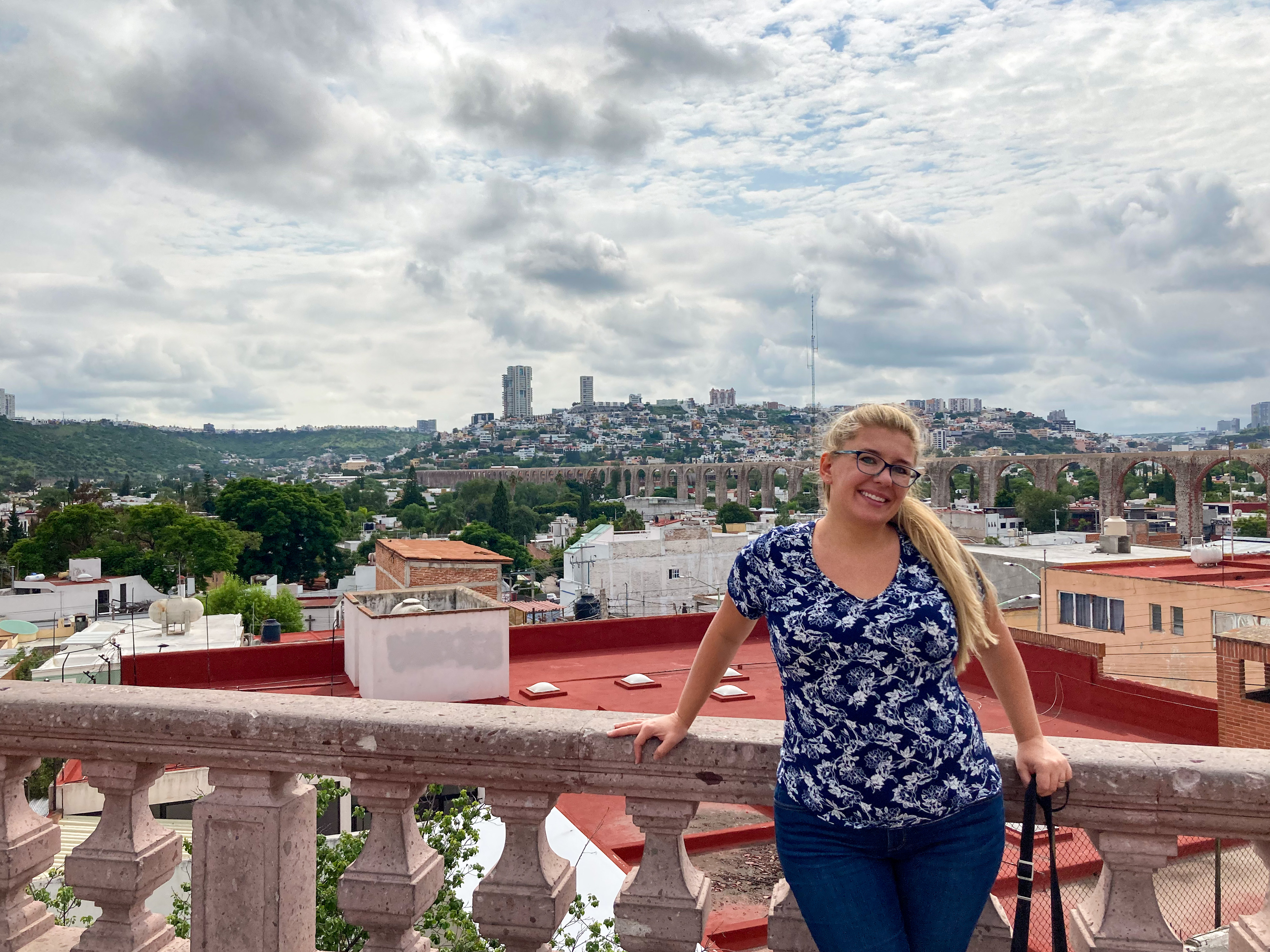 Your (somewhat) fearless author, posing in front of the Aqueduct (far background). A smile is on her face! She is wearing a blue and white patterned shirt and dark blue jeans and has blonde hair and glasses. The colors in the buildings are pinks and reds. The sky is cloudy with only the merest hints of blue.