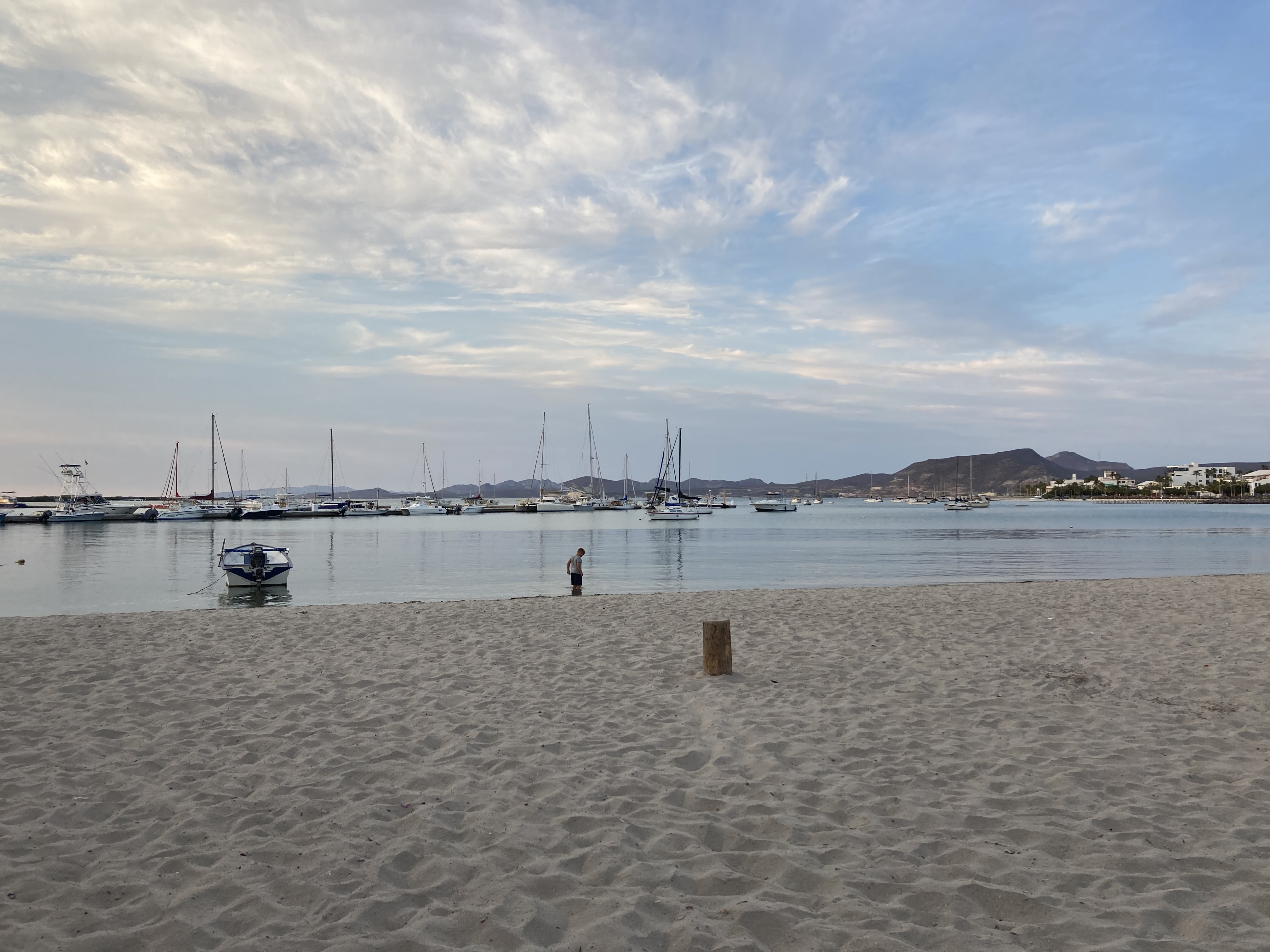 A photo of a beach, the sand taking up the front part of the photo. There is a child at the shoreline looking down. There are boats scattered around the background. The sky is blue with the clouds looking like a painting, but it's real life.