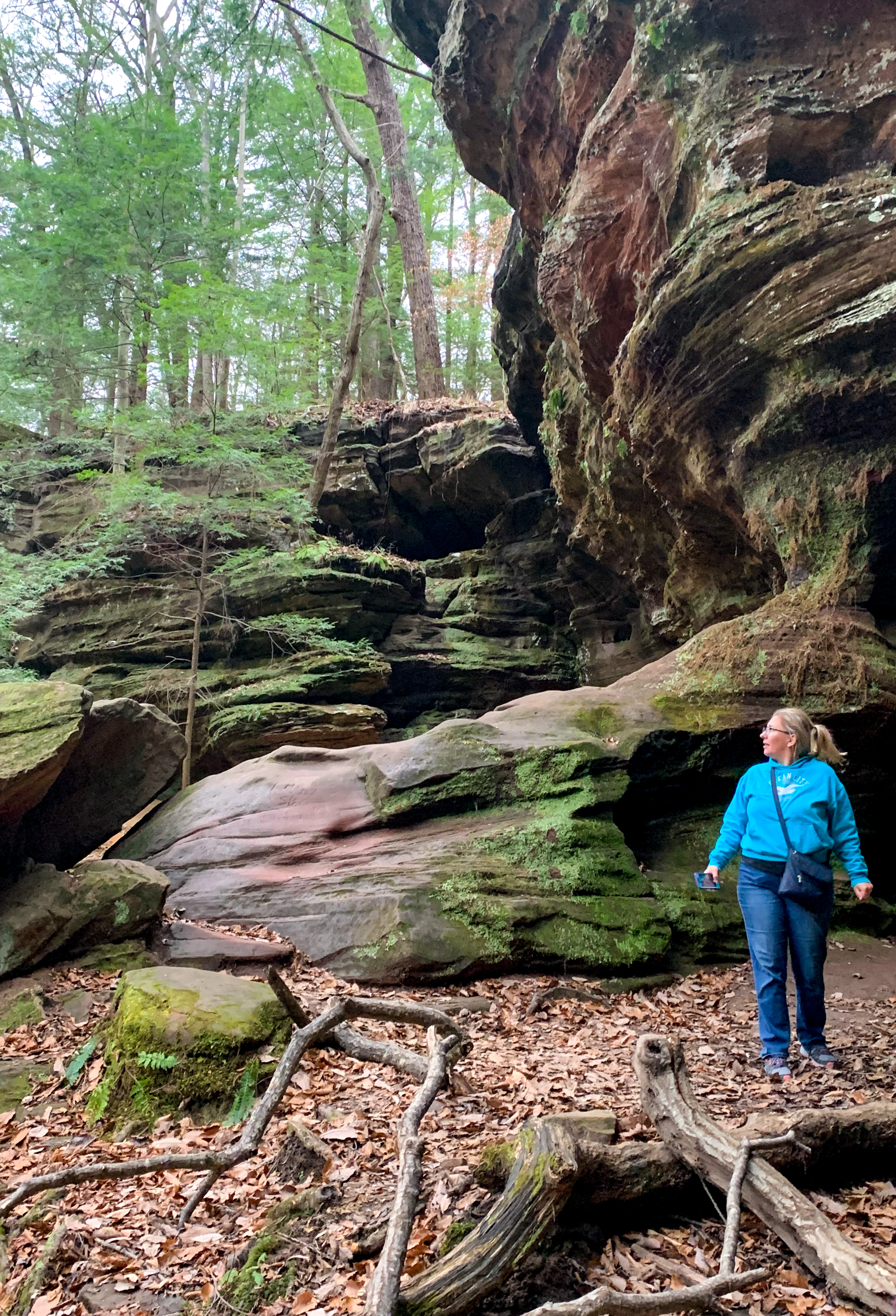 A blonde woman wearing a teal sweatshirt, jeans, and glasses looks up and to the left of the photo from her spot on the bottom right. Behind her, the cliff is showing off its rocky sections.