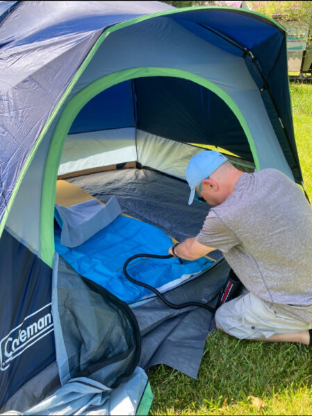 Man in a blue hat and gray shirt is using a device to blow up a single person camping mat that's already inside a blue and green tent.