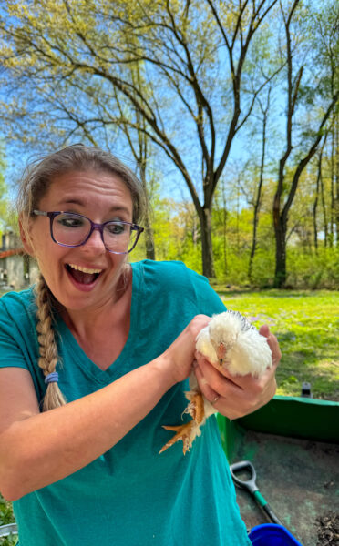 A woman with her hair in a braid and a teal shirt, holds a large, white feathered chick.