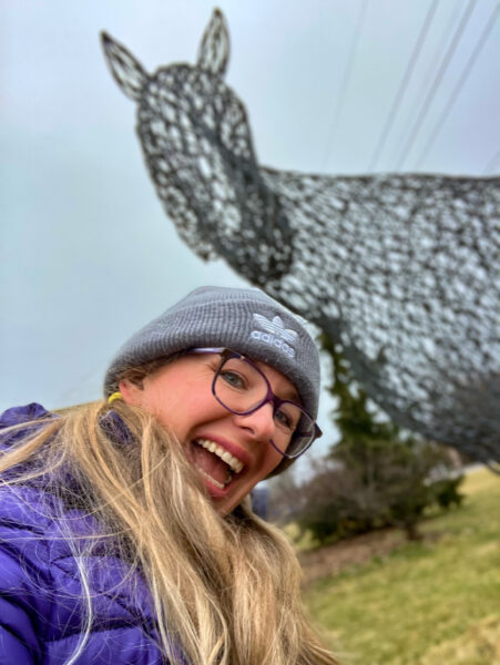 A huge wire horse art installation is behind a woman in a gray hat and purple coat, who is wearing purple rimmed glasses.