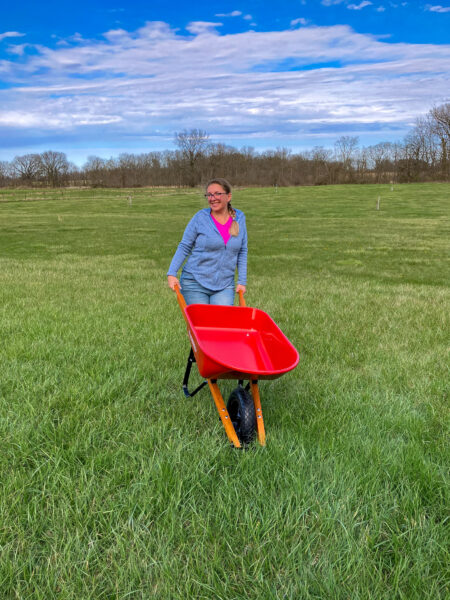 Brown haired woman in blue zip up, stands holding a red wheel barrow by its wooden handles. Grass, sky, and trees are in the background.