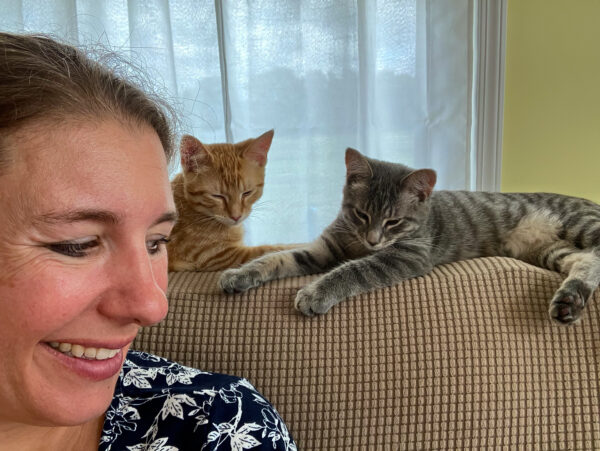 A woman smiles with her head turned slightly away from the camera. Behind her, on a couch back, are two kittens. One is an orange striped cat and the other is gray striped.