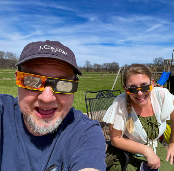 A man in a hat and solar eclipse glasses is in the foreground and a woman with brownish hair and solar eclipse glasses is in th ebackground. Both are smiling.